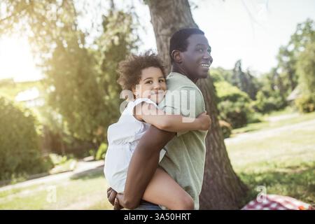 Der lächelnde Schwarze Vater trägt seine Tochter auf dem Rücken und genießt einen sonnigen Tag im Park. Ideal für Familienleben, Eltern und Outdoor-Spaß. Stockfoto