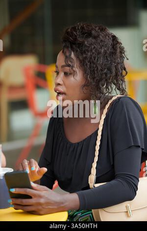 Emotionale und stilvolle Schwarze Frau an einem Café-Tisch spricht auf ihrem Smartphone mit Freunden in der Nähe. Das städtische Coffeeshop stellt die soziale Interaktion und das Stadtleben in Szene. Ideal für Lifestyle-Kampagnen, Kommunikationsthemen oder Social-Media-Inhalte. Stockfoto