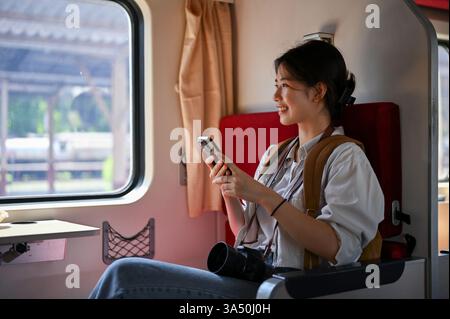 Lächelnde asiatische Reisende sitzt in einem Zug, hält ein Smartphone und blickt aus dem Fenster. Geeignet für Reisen, mobile Technologie und Reisebilder. Ein helles, optimiertes Aktienbild mit einem Sinn für Erkundung. Stockfoto
