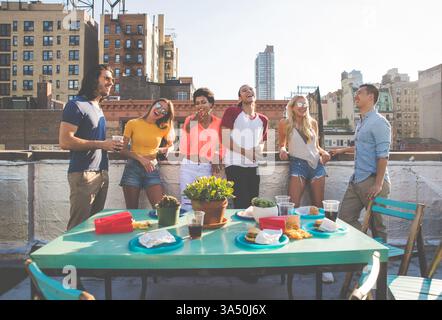 Eine fröhliche Gruppe verschiedener Freunde steht auf einem sonnendurchfluteten Dach, lehnt sich auf das Geländer und teilt ein Lächeln. Ideal für Lifestyle, Freundschaft und soziale Kampagnen, um Glück, Zweisamkeit und urbanes Leben im Freien festzuhalten. Stockfoto