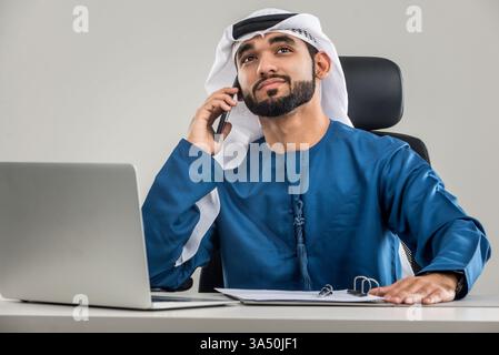 Ein Mann aus dem Nahen Osten, der wegschaut, während er in einem modernen Büro telefoniert. Er sitzt mit einem Laptop in der Nähe und vermittelt Geschäftsfokus und eine moderne Arbeitsumgebung. Hervorragend geeignet für Unternehmenskultur, regionale Geschäfte und Visuals von Bürotechnik. Stockfoto