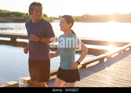 Ein lächelndes chinesisches Paar in den 50ern joggen bei Sonnenuntergang gemeinsam durch einen Park am See. Die beleuchtete Szene unterstreicht die Vitalität und Zweisamkeit während des Fitnessstudios im Freien. Dieses Image eignet sich gut für Pensionierung, gesunde Lebensweise und Familien-Wellness-Kampagnen. Stockfoto