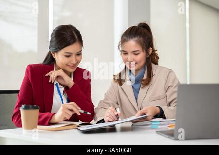 Zwei asiatische Geschäftsfrauen diskutieren in einem modernen Büro über Arbeit und prüfen Finanzberichte. Ideal für Unternehmenskommunikation, Teamarbeit und professionelle Zusammenarbeit im Bereich Finanzdienstleistungen. Eine klare, dynamische Szene, in der Kollegen an einem Projekt zusammenarbeiten. Stockfoto