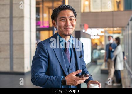 Lächelnder asiatischer Geschäftsmann in blauem Anzug steht mit Smartphone und Kaffeetasse in einem modernen Bürogebäude. Dieses professionelle Porträt unterstützt Führungsqualitäten, Netzwerke und Vertrauen am Arbeitsplatz in einer städtischen Umgebung. Stockfoto
