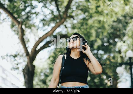 Die lächelnde hispanische Frau mit Kopfhörern steht tagsüber in einem sonnendurchfluteten Park. Die Outdoor-Lifestyle-Szene fängt einen entspannten Moment der Musik inmitten von Bäumen ein, die auf Frühlings- oder Sommerstimmung hindeutet. Ein helles, ansprechbares Image für Wellness-, Freizeit- und Unterhaltungskampagnen. Stockfoto
