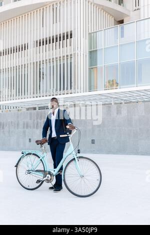 Fröhlicher schwarzer Angestellter in formellem Anzug steht neben einem Fahrrad vor einem Bürogebäude aus Glas und Beton. Die urbane Kulisse vermittelt professionelles, modernes Stadtleben und Pendlerstil. Ideal für Firmenporträts, Business Lifestyle und Startup-Marketing. Stockfoto