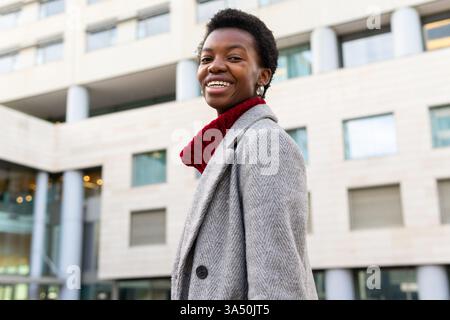 Seitenansicht einer lächelnden jungen Afroamerikanerin auf einer Stadtstraße. Urbanes Lifestyle-Bild, das Selbstvertrauen, Stil und alltägliche Momente vermittelt. Stockfoto