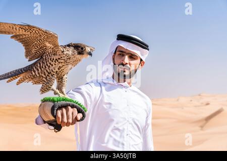Porträt eines Mannes aus dem Nahen Osten, der in der Wüste einen traditionellen Kandura trägt und einen Falken hält. Ein beeindruckendes Bild, das Falknerei, Erbe und arabische Kultur vor Sanddünen hervorhebt. Geeignet für Reisen, Kultur und Business Lifestyle Themen. Stockfoto