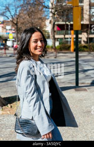 Seitenansicht einer lächelnden jungen hispanischen Frau, die an einem sonnigen Tag in der Stadt spaziert, mit einem blauen Himmel und verschwommenen Gebäuden und Bäumen im Hintergrund. Ideal für den urbanen Lebensstil und alltägliche Freizeitaktivitäten. Stockfoto
