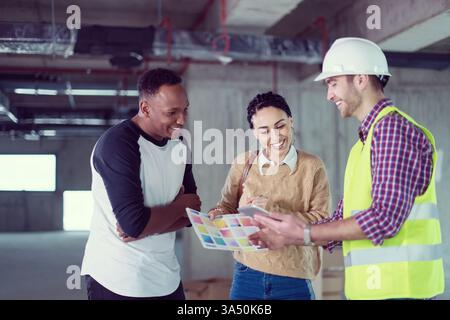 Ein Architekt zeigt einem jungen multiethnischen Paar auf einer Baustelle auf einem Tablet-PC die Pläne eines Hauses. Das Bild vermittelt Zusammenarbeit, modernes Design und Kundenbindung beim Hausbau. Ideal für Immobilien-, Architektur- und Baumarkenwerbung. Stockfoto