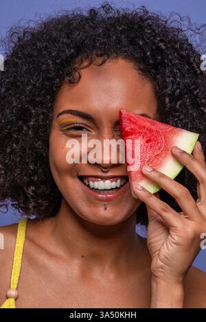 Eine freundliche, charismatische afroamerikanische Frau lächelt in die Kamera und versteckt ein Auge hinter einem Stück Wassermelone. Ein helles, sommerliches Porträt mit Früchten und Lifestyle-Vibes. Geeignet für Lifestyle-, Food- und Schönheitskampagnen. Stockfoto