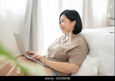 Seitenansicht einer großen asiatischen Frau, die einen Laptop in ihrem Schlafzimmer benutzt. Eine gemütliche Arbeit aus der Heimszene, die sich für freiberufliche Mitarbeiter, Online-Schulungen und Home-Office-Themen eignet. Stockfoto