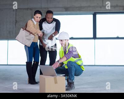 Verschiedene Gruppen stehen mit einem Tablet, während ein kaukasischer Ingenieur sich hockt, um die Pläne für die Hausgestaltung zu besprechen. Ein Bild mit dem Thema Bau und Architektur, das sich für Wohnbauprojekte eignet. Stockfoto