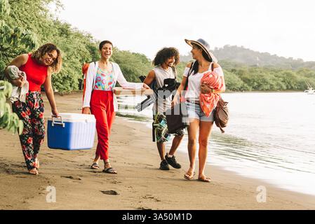 Fröhliche hispanische Freunde genießen ein sonniges Picknick am Strand und spazieren zusammen mit Speisen und Vorräten. Die lebhafte Küstenlandschaft bietet Freundschaft, Freizeit im Freien und eine entspannte Sommeratmosphäre, ideal für Reisen, Lifestyle und Urlaubsbilder. Stockfoto