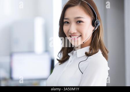 Eine fröhliche chinesische Geschäftsfrau mit Headset sitzt an einem Tisch in einem hellen Büro. Dieses Porträt unterstreicht den vertrauenswürdigen Kundenservice und die Kommunikation am Telefon. Das Headset und das Büro vermitteln eine Callcenter-Atmosphäre. Ideal für Business-, Tech-Support- und Office-Workflow-Kampagnen. Stockfoto