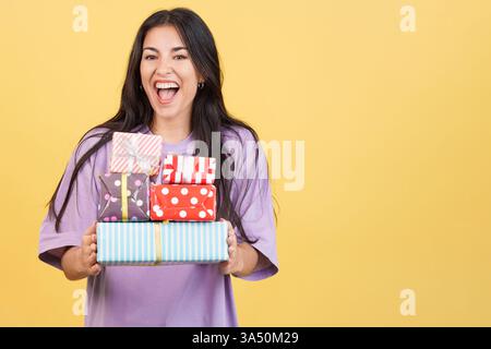 Fröhliche weiße Frau hält einen Stapel bunter Geschenke in einem hellen Atelier vor gelbem Hintergrund. Diese festliche Szene ist ideal für Feiertags- und Weihnachtskampagnen, Geschenke und Werbematerial. Stockfoto