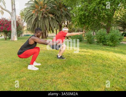 Durchgehende Seitenansicht verschiedener männlicher Athleten in Fitnesskleidung, die Squats auf einem grasbewachsenen Rasen mit Palmen durchführen. Diese energiegeladene Outdoor-Szene unterstreicht Kraft, Training und Ausdauer. Ideal für Fitness- und Wellnesskampagnen, Park-Workouts und Outdoor-Sportarten. Stockfoto