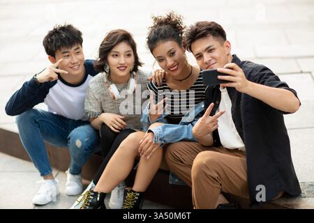 Eine lächelnde Gruppe chinesischer Freunde macht tagsüber ein Selfie im Freien auf dem Bürgersteig. Ungezwungene urbane Atmosphäre mit Smartphones und sozialen Verbindungen für Jugendkultur und Stadtleben. Ideal für Lifestyle-, Reise- und Gruppenaktivitäten, bei denen die Zweisamkeit betont wird. Stockfoto