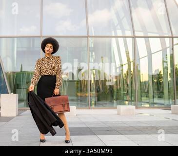 Hispanische Geschäftsfrau mit lockigem Haar, die neben einem Glashaus steht und eine Jacke hält. Die professionelle Pose vermittelt Selbstvertrauen, Stil und das Arbeitsleben in der Innenstadt. Geeignet für Unternehmens-, Karriere- und moderne Bürobilder. Stockfoto