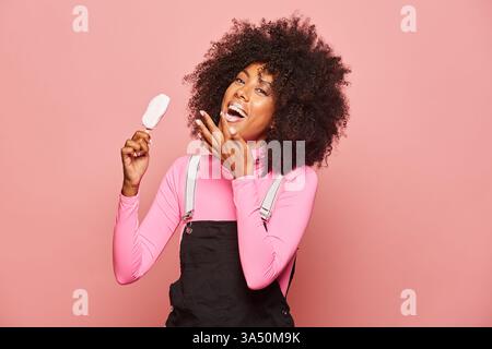 Fröhliche junge afroamerikanische Frau genießt ein Eis in einer zwanglosen Umgebung. Sie lächelt vor einem rosa Hintergrund und fängt sommerliche Erfrischungen und Glücksgefühle ein. Ideal für Marketingkampagnen mit abwechslungsreichen, fröhlichen Frauen und angenehmen Sommerspezialitäten. Stockfoto