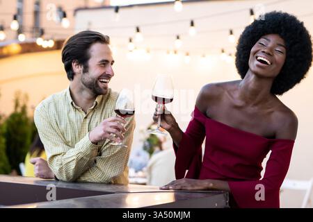 Verschiedene Gruppen auf einem Dach, bei denen ein Wiedersehen mit Freunden und Familie unter Lichterketten gefeiert wird. Ideal für Lifestyle- und Eventkampagnen, bietet er warme Zweisamkeit, abendliche Atmosphäre und geselliges Essen im Freien. Stockfoto