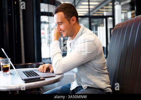 Ein lächelnder hispanischer Mann sitzt an einem Café-Tisch und legt seinen Kopf auf seine Hand, während er an einem Laptop arbeitet. Tagsüber vermittelt die Umgebung legere Geschäftsabläufe und modernen Arbeitsstil, ideal für Cafés oder Remote-Arbeiten. Stockfoto