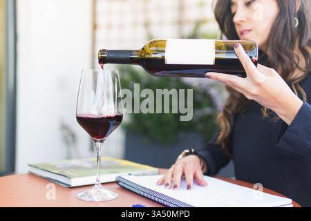 Hispanische Sommelierin saß an einem Tageslichttisch und goss Rotwein in einen Becher. Ideal für Weinproben, Speisen und Restaurants im Freien oder Weinkellereien. Die Umgebung verbindet Gourmet-Ambiente mit einer luftigen, sonnendurchfluteten Atmosphäre. Stockfoto