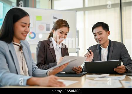 Lächelnder asiatischer Geschäftsmann, der mit weiblichen Kollegen zusammenarbeitet, die am Tisch sitzen und Finanzberichte im Büro diskutieren Stockfoto