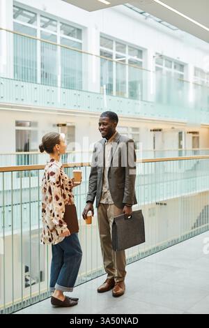 Lächelnder schwarzer Geschäftsmann mit Brille, der Kaffee und Tasche hält und mit einer kaukasischen Kollegin plaudert, während er im Büro auf dem Balkon steht... Stockfoto
