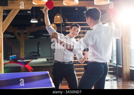 Junge chinesische Geschäftsleute spielen Tischtennis in einem modernen Büro. Der energiegeladene Moment mit Anfeuern und Teamwork macht dieses Image für Corporate Branding, Bürokultur und sportliche Kampagnen geeignet. Stockfoto