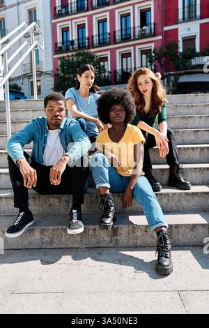 Ernsthafte, vielfältige Gruppe von Freunden, die auf einer Außentreppe in einer sonnigen Stadtszene sitzen. Dieser multiethnische, urbane Moment ist ideal für Lifestyle, Freundschaft und Teamwork mit einer modernen Atmosphäre. Stockfoto
