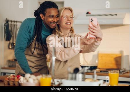 Fröhliches multikulturelles Paar zu Hause teilen sich eine Telefonkonferenz am Telefon, während sie eine Mahlzeit in der Küche zubereiten. Das Bild hebt die Verbindung, moderne Technologie und den Familienalltag hervor. Es eignet sich gut für Materialien über Fernarbeit, Online-Kommunikation und multikulturelle Haushalte. Stockfoto