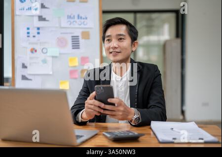 Ein lächelnder asiatischer Geschäftsmann schaut weg, während er ein Smartphone an seinem Schreibtisch im Büro hält. Dieses zeitgenössische Porträt zeigt einen technisch versierten Profi in einer Unternehmensumgebung. Geeignet für Geschäftskommunikation, Mobiltechnologie und südostasiatische Märkte. Stockfoto