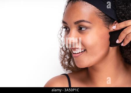 Optimistische junge hispanische Frau mit lockigem Haar lächelt während einer Gua-Sha-Gesichtsmassage. Die weiße Kulisse macht dieses Schönheits- und Hautpflegebild ideal für Wellness-Kampagnen. Stockfoto