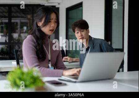 Asiatischer männlicher Büroangestellter, der versucht, gestressten weiblichen Kollegen mit Laptopproblemen zu helfen, die an Schreibtischen im Büro sitzen Stockfoto