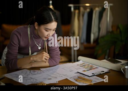 Professionelle asiatische Modedesignerin skizziert neue Kostümideen in ihrem Atelier. Dieses fokussierte Porträt hebt Kreativität, Designarbeit und Bekleidungsherstellung für Kampagnen der Modeindustrie hervor. Stockfoto
