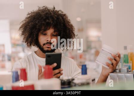 Ein multirassischer Mann mit Afrobart überprüft Medikamenteninformationen auf seinem Smartphone neben Apothekenregalen. Ideal für Themen wie Online-Apotheke, Medikamentenberatung und Gesundheitseinkäufe. Stockfoto