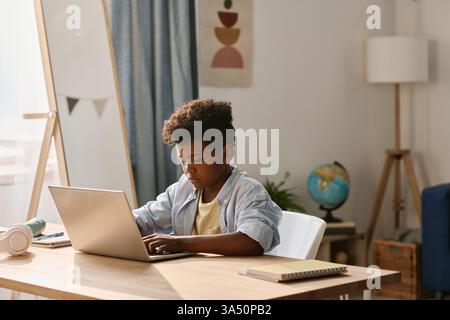 Afrikanischer Junge, der auf einem Laptop schreibt, um online Hausaufgaben zu machen, während er an einem Schreibtisch in seinem Zimmer sitzt. Eine fokussierte Indoor-Studienszene, die sich gut für Themen wie Bildung, E-Learning und Fernunterricht eignet. Geeignet für Inhalte über Technologie im Haushalt und digitale Bildung. Stockfoto