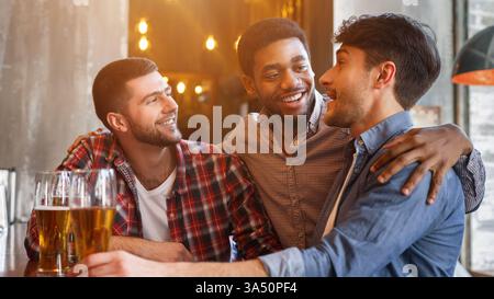 Ein lächelnder Schwarzer und kaukasische Freunde unterhalten sich, während sie zusammen am Tisch sitzen und ein Glas Bier in der Bar trinken Stockfoto