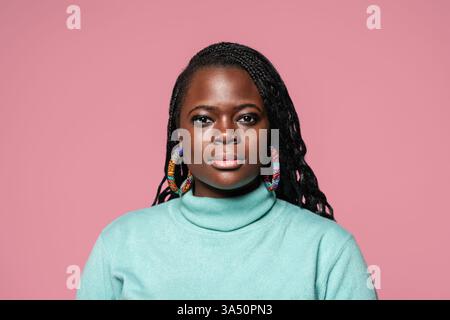 Nahporträt einer Afrikanerin mit geflochtenen Haaren mit türkisfarbenem Rollkragen und farbenfrohen Ohrringen. Sie steht vor einem rosafarbenen Hintergrund und schafft ein lebendiges, modefokussiertes Bild. Die Aufnahme betont Schönheit und Selbstvertrauen mit einem modernen Studio-Feeling. Stockfoto