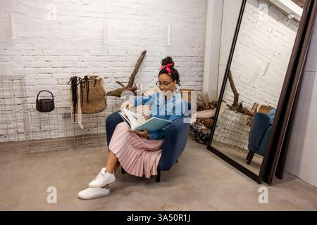 Die lächelnde schwarze Frau im Boho-Stil sitzt auf einem Sofa in einem hellen Kunststudio und liest eine Zeitschrift. Sie trägt eine Brille und ein rotes Haarband, was einen stilvollen, kreativen Look schafft. Dieses Bild eignet sich gut für Kunststudios, Heimwerkergewerbe und Unternehmerinnen. Stockfoto