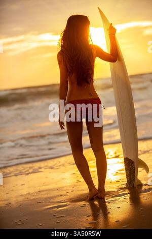 Asiatische Surferin im Bikini steht mit ihrem Surfbrett am Strand bei Sonnenuntergang. Sie blickt entlang der Küste, während warmes Licht eine tropische Stimmung erzeugt. Ideal für Reisen, Sport und Lifestyle an der Küste. Stockfoto