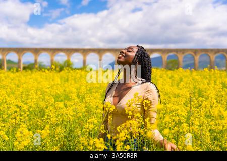 Porträt einer schwarzen Frau mit Zöpfen, die auf einer Wiese aus gelben Blumen unter natürlichem Sonnenschein stehen. Ideal für Lifestyle-, Natur- und Outdoor-Themen. Stockfoto