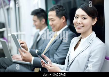 Chinesische Geschäftsfrau mit Kollegen, die Gadgets in einer U-Bahn nutzen und damit eine vernetzte, mobile Arbeitskultur widerspiegeln. Geeignet für Themen wie Pendeln in der Stadt, Teamarbeit und Technologienutzung im Transit. Ein dynamischer Moment des Stadtlebens auf Geschäftsreisen. Stockfoto
