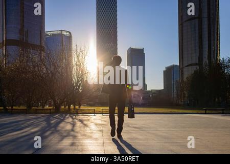 Chinesischer Geschäftsmann, der in einer Stadt spaziert, während er mit dem Handy spricht. Diese lange, hintergrundbeleuchtete urbane Szene fängt einen modernen Profi inmitten hoher Wolkenkratzer ein. Stockfoto