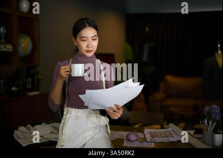 Asiatische Modedesignerin, die sich auf die Millennials konzentriert, trinkt Kaffee und überprüft Modellskizzen in einem Studio. Dieser stilvolle Profi strahlt Kreativität und Unternehmergeist aus und vereint Stoffe und Muster. Ideal für Modedesign, Boutique-Branding und Startup-Kampagnen. Stockfoto