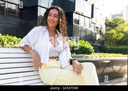 Glückliche junge hispanische Frau sitzt auf einer Holzbank in einem sonnigen Park und lächelt, während sie wegschaut. Sie trägt ein stilvolles, lässiges Outfit neben üppigen Büschen an einem hellen Tag. Perfekt für Lifestyle-, Park- und Outdoor-Freizeitbilder und Reiseangebote. Stockfoto