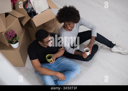 Fröhliches afroamerikanisches Paar, das sich in ihrem neuen Zuhause entspannt und auf dem Boden mit Kaffee inmitten ausgepackter Kisten sitzt. Ein warmer Moment, der Bewegung, Eingewöhnung und den Alltag zu Hause vermittelt. Ideal für Immobilien-, Umzugsaktionen und Lifestyle-Kampagnen für Familien. Stockfoto