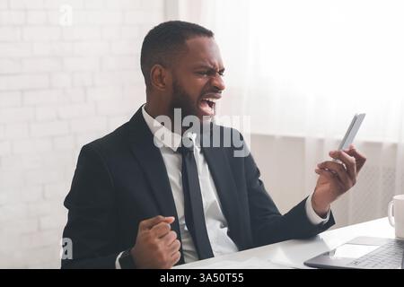 Schwarzer Geschäftsmann mit wütendem Ausdruck, der auf ein Smartphone am Schreibtisch mit Laptop im Büro blickt Stockfoto
