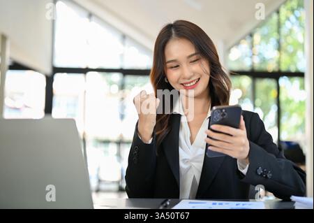 Charmante junge asiatische Geschäftsfrau reagiert auf gute Nachrichten auf ihrem Smartphone im Büro. Eine lebendige Arbeitsplatzszene, in der mobile Kommunikation, Geschäftserfolg und weibliche Führungsqualitäten hervorgehoben werden. Stockfoto